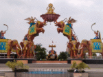 Grand decorative entrance featuring giant elephant statues, winged figures, and a goddess seated on a lotus above the arch under a cloudy sky.