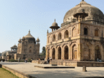 Ancient Mughal-style tomb architecture with large domes, sandstone carvings, arched corridors, and open courtyard under clear blue sky.