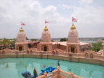 Beautiful Hindu temple complex with three pink temple towers, red flags, and a large water pond under a cloudy sky.