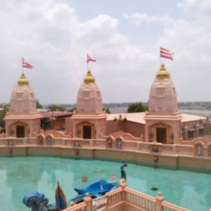 Beautiful Hindu temple complex with three pink temple towers, red flags, and a large water pond under a cloudy sky.