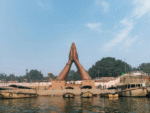 Large folded-hands sculpture at the riverside ghat with boats floating on the water and a clear blue sky in the background.