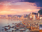 Sunrise view of Varanasi ghats on the banks of the Ganga River with colorful boats, crowded temples, historic buildings, and devotees gathered along the riverside.